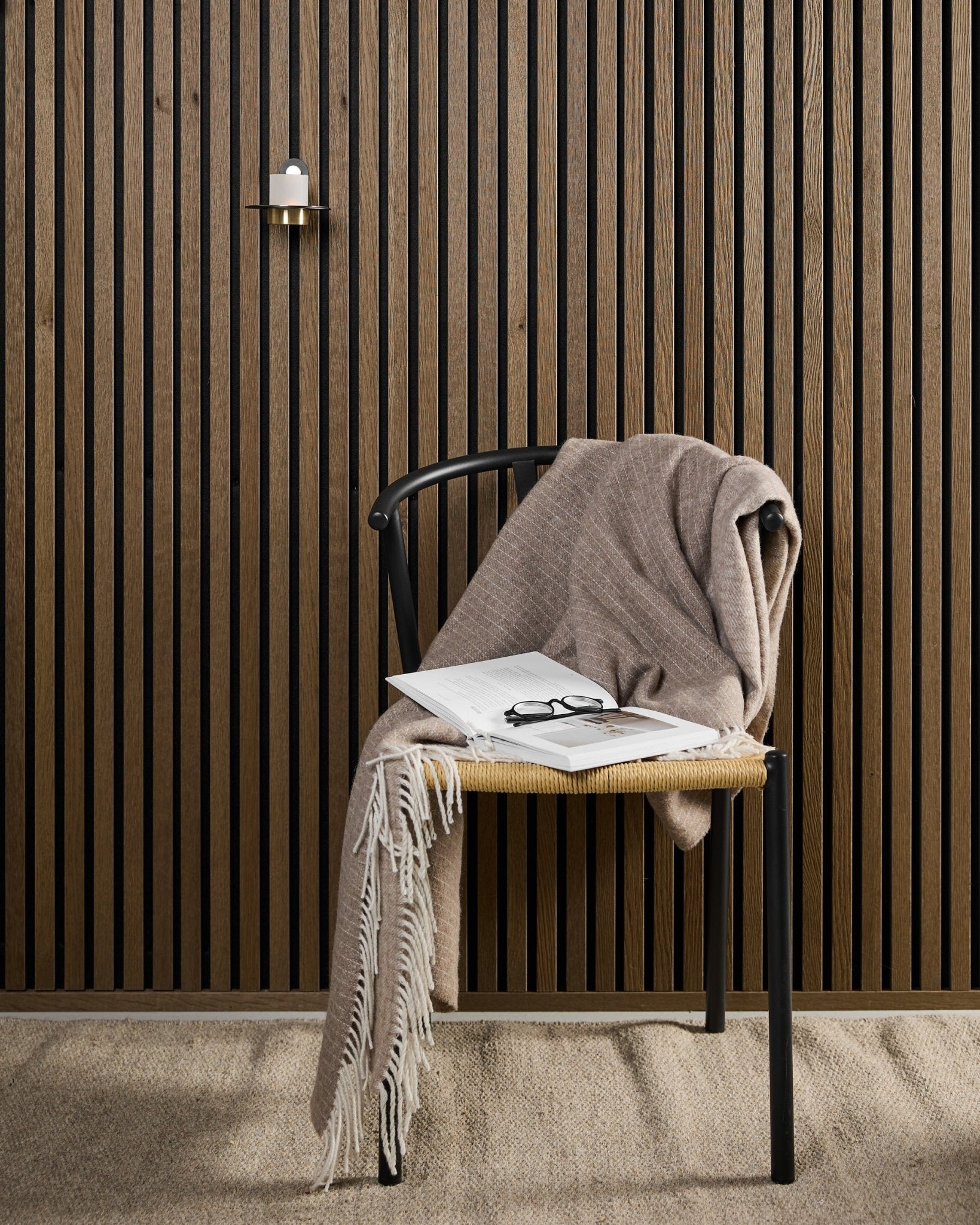 Een moderne stoel met een beige plaid, een open boek en een bril staat naast een Akupanel akoestisch houten paneel en een black en brass WoodUpp Norr kaarsenhouder.